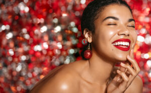A woman with glowing skin and vibrant red lipstick smiles with her eyes closed against a festive, glittering red and silver bokeh background. She is wearing a large red bauble earring and has her hand gently touching her chin.