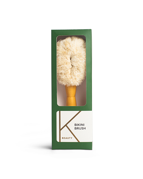 Image of a product package showing a "Bikini Brush." The brush has a wooden handle and light-colored bristles, displayed in a rectangular green box with a transparent front. The label on the lower right corner reads "K Beauty" and "Bikini Brush.