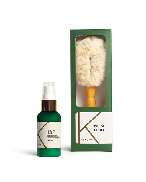 A green bottle of Bikini Balm with a white label featuring the letter "K" and a green rectangular box containing a fluffy brush, also labeled with "Bikini Brush" and "K Beauty." The brush has a natural wooden handle.