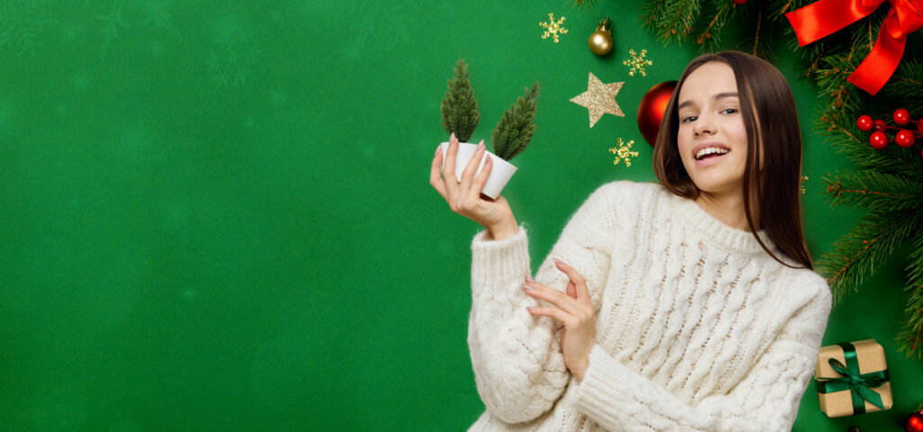 A smiling woman in a white sweater holding a small potted plant with two miniature trees in front of a festive green background decorated with holiday ornaments, stars, and ribbons.